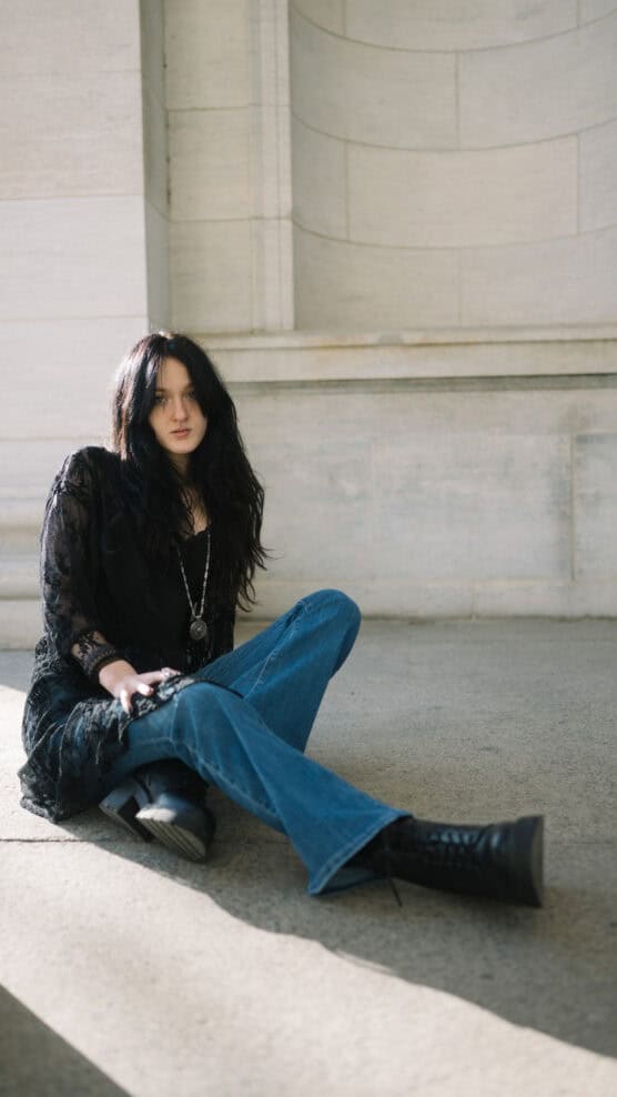 A person with long black hair, wearing a black lace top, blue jeans, and black boots, sits on the ground against a stone wall in natural light, looking toward the camera. by Justin Salem Meyer