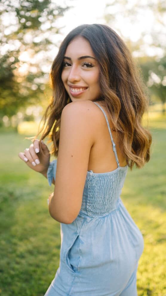 A young woman with wavy brown hair in a light blue sundress smiles over her shoulder while standing outside in a sunlit, grassy park. by Justin Salem Meyer