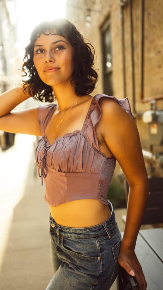 A woman with short curly hair wearing a purple ruffled crop top and jeans stands outside in sunlight, smiling slightly with one hand touching her hair. The background shows a brick building and an alleyway. by Justin Salem Meyer