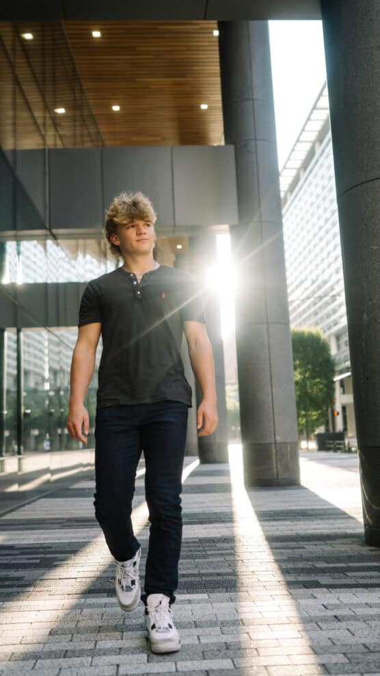 A young man with blond hair walks confidently on a city sidewalk, wearing a black polo shirt, dark jeans, and white sneakers. Sunlight shines between buildings, creating a bright glare behind him. by Justin Salem Meyer