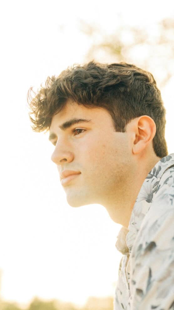 A young man with short, dark, wavy hair gazes thoughtfully into the distance. He is wearing a light-colored, patterned shirt, and the background is softly lit with a warm, natural light. by Justin Salem Meyer