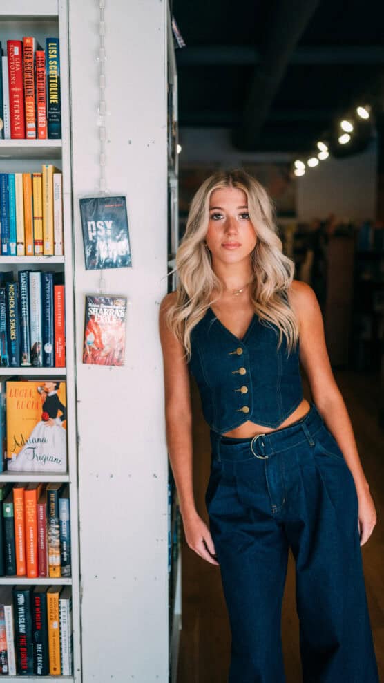 A young woman with long blonde hair, wearing a sleeveless denim top and matching high-waisted pants, stands next to a bookshelf in a bookstore, looking directly at the camera. by Justin Salem Meyer