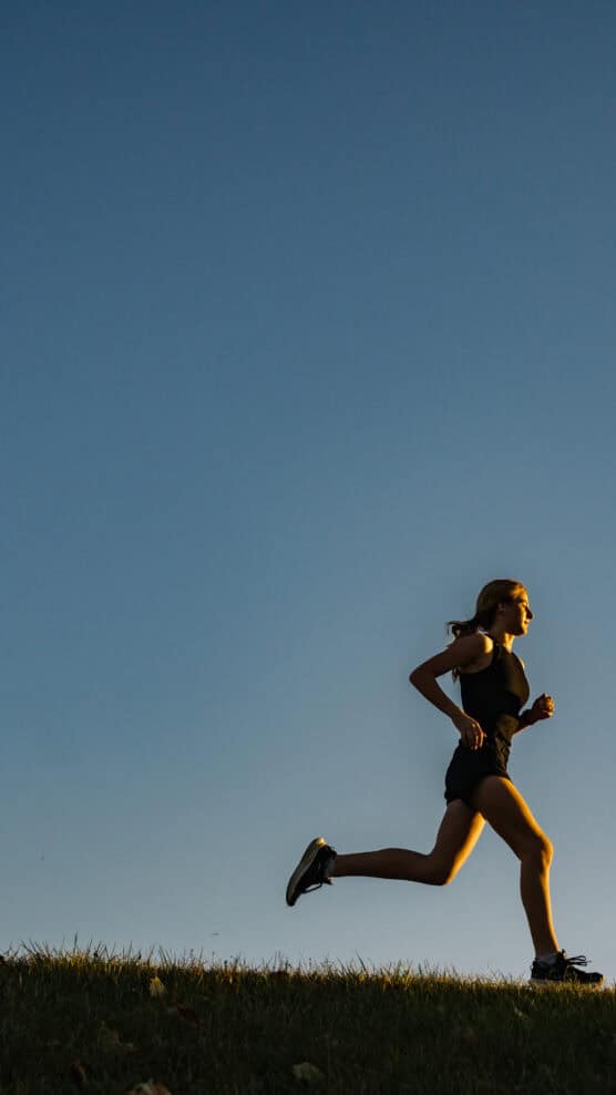 A person with a ponytail runs outdoors on grass under a clear blue sky, wearing athletic clothes and running shoes, with sunlight highlighting their profile. by Justin Salem Meyer