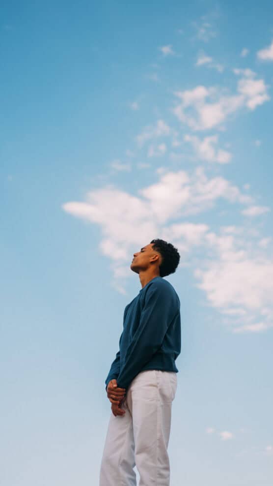 A person wearing a blue long-sleeve shirt and white pants stands outdoors, looking up at the sky with light clouds scattered across a blue background. by Justin Salem Meyer