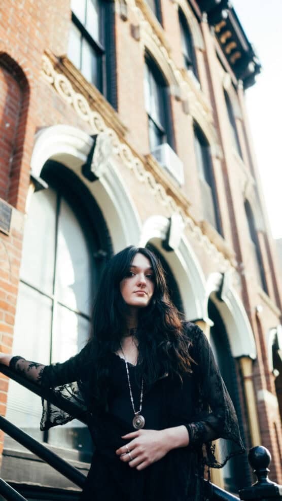 A woman with long dark hair wearing a black outfit and pendant necklace stands on a staircase outside a brick building with large arched windows, looking confidently at the camera. by Justin Salem Meyer