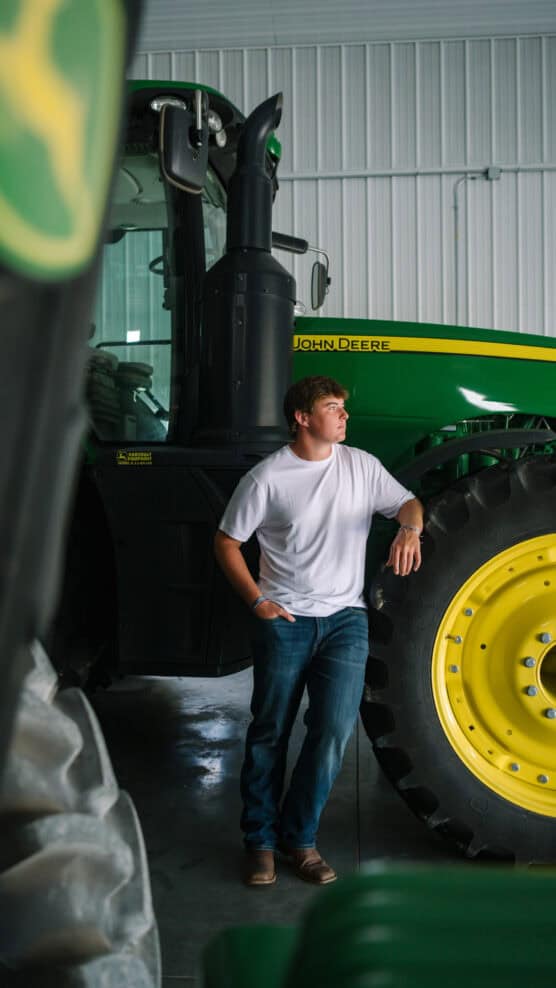 A young man in a white t-shirt and jeans leans against a large green and yellow John Deere tractor inside a well-lit metal garage or barn. by Justin Salem Meyer