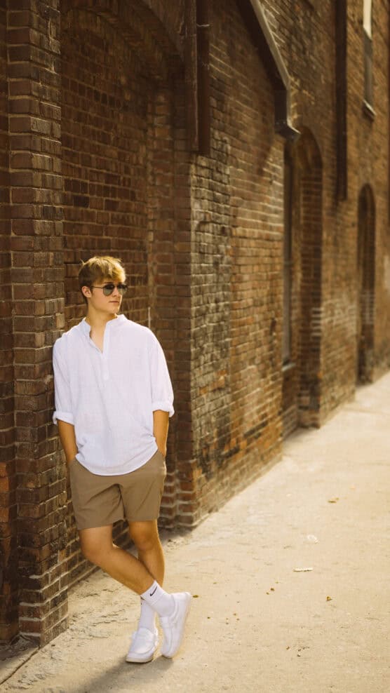 A young person in sunglasses, a white shirt, and tan shorts leans against an old brick wall with hands in pockets, looking relaxed. The background features more brick archways and a sunlit sidewalk. by Justin Salem Meyer