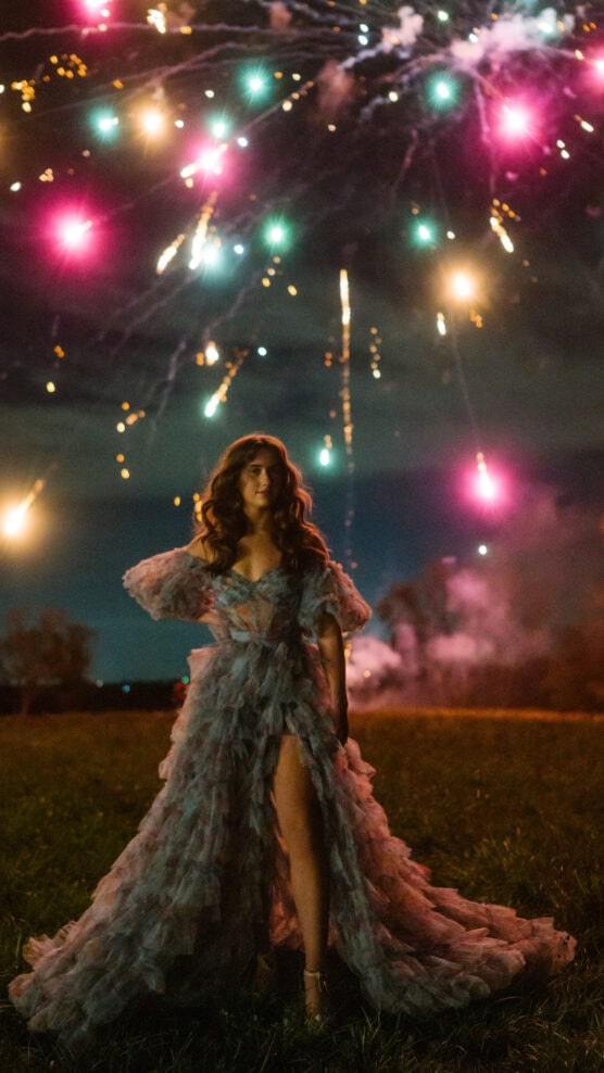 A woman in a dramatic, ruffled gown stands outdoors at night under a sky filled with colorful fireworks, with smoke and glowing lights illuminating the scene. by Justin Salem Meyer