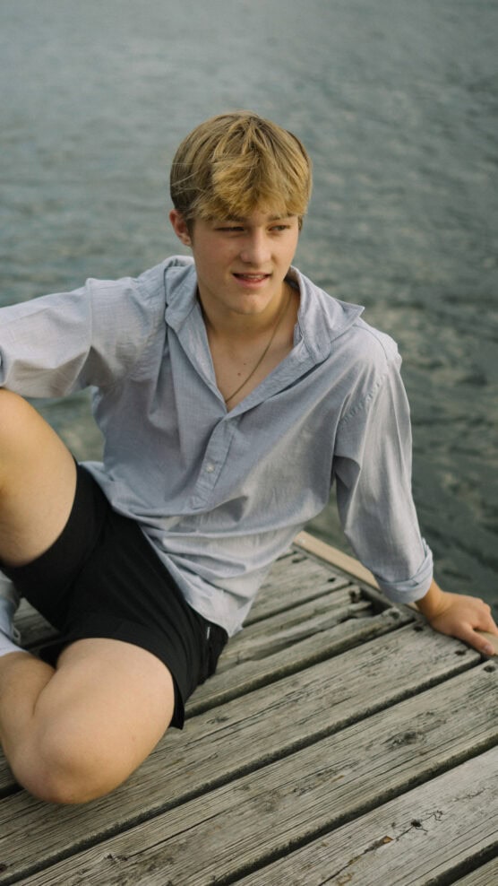 A young person with blonde hair sits on a wooden dock by the water, wearing a light blue button-up shirt and black shorts, looking off to the side with a relaxed expression. by Justin Salem Meyer