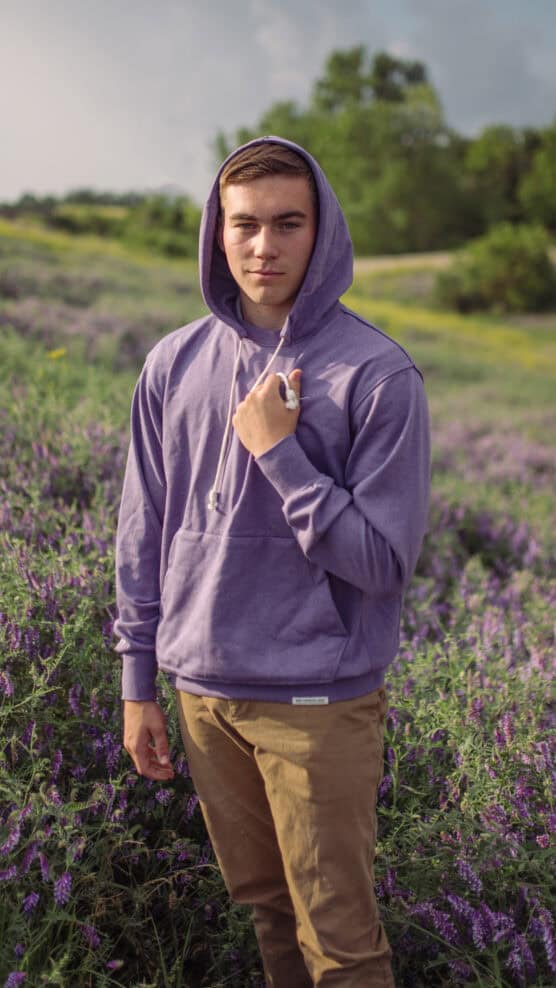 A young man wearing a purple hoodie and khaki pants stands in a field of purple wildflowers, holding a small white flower in his left hand. Green trees and cloudy sky are visible in the background. by Justin Salem Meyer