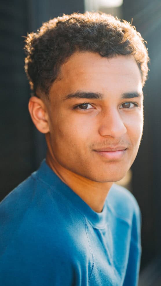A young man with short curly hair wears a blue shirt and looks slightly over his shoulder, smiling softly at the camera. Sunlight highlights his face and the background is softly blurred. by Justin Salem Meyer