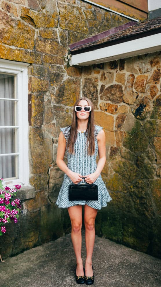 A woman stands outdoors against a stone wall, wearing a short blue patterned dress, black handbag, black shoes, and white sunglasses. A window with curtains and pink flowers is visible to the left. by Justin Salem Meyer
