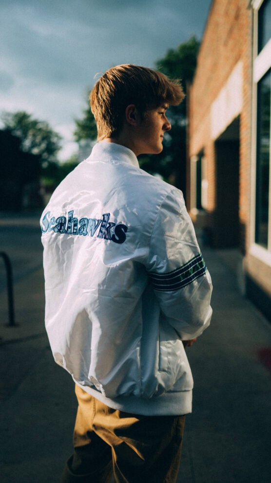 A young man stands outdoors, wearing a white Seahawks jacket and brown pants. He is looking to his left, and sunlight highlights his hair and jacket. A brick building and some trees are in the background. by Justin Salem Meyer