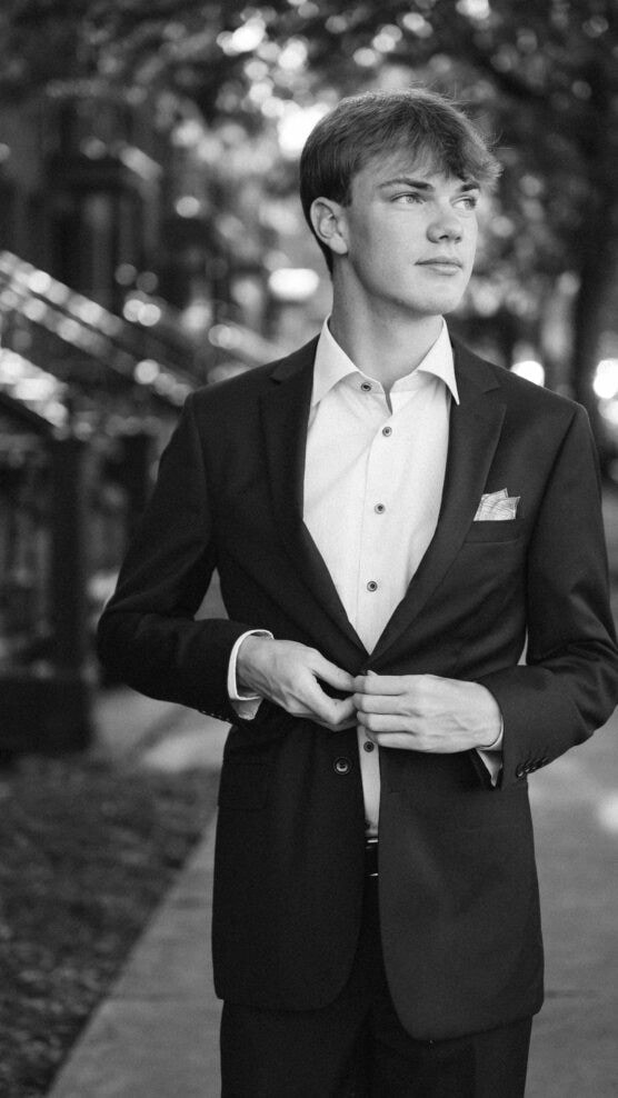 A young man in a suit stands on a sidewalk in a city, adjusting his jacket. The image is black and white, with trees and buildings blurred in the background. He looks off to the side with a thoughtful expression. by Justin Salem Meyer