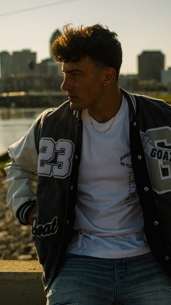 A young man with short dark hair, wearing a varsity jacket and white t-shirt, sits outdoors in front of a river with a city skyline in the background, looking to his left. The lighting is warm and golden. by Justin Salem Meyer