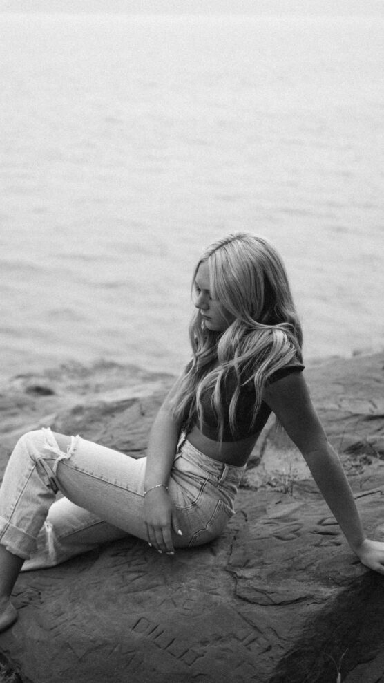 Black and white photo of a woman with long hair sitting on a rocky surface by the water, looking down, with one arm resting on the ground and the other on her knee, wearing a crop top and jeans. by Justin Salem Meyer