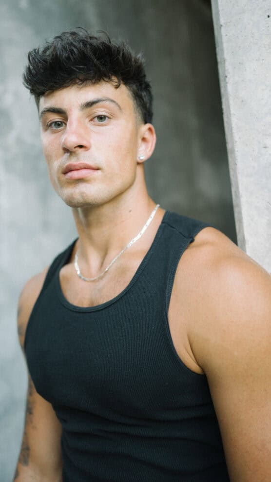 A young man with short dark hair wearing a black tank top and a silver necklace stands against a concrete wall, looking confidently at the camera. by Justin Salem Meyer
