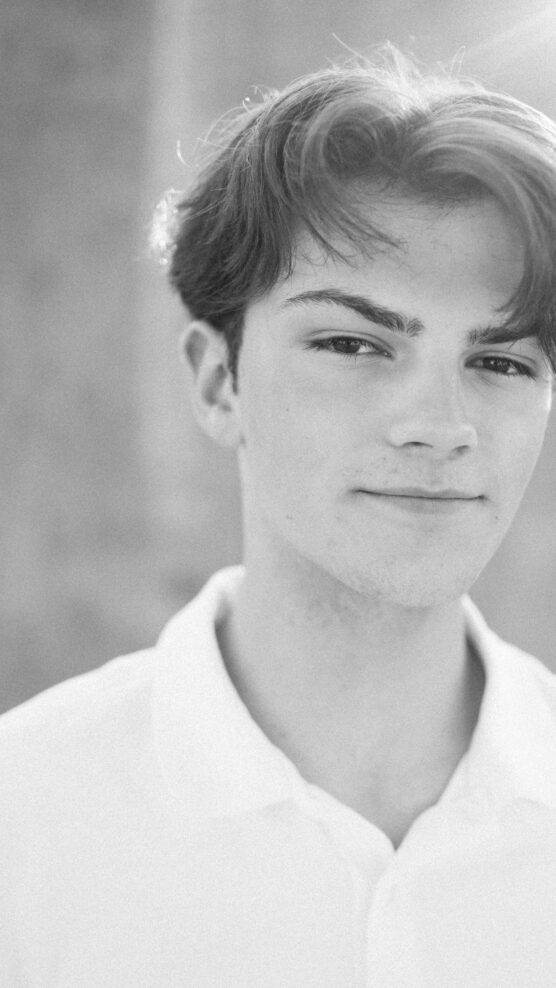 Black and white portrait of a young person with short, tousled hair wearing a white collared shirt, softly smiling and looking into the camera with a blurred background. by Justin Salem Meyer