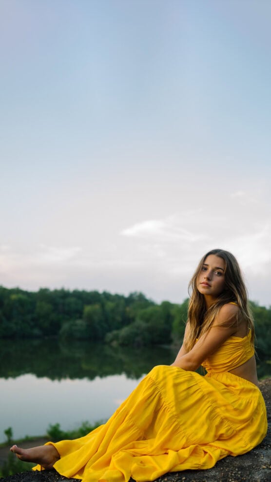 A woman in a yellow dress sits by a calm lake with green trees in the background, looking thoughtfully at the camera under a clear sky. by Justin Salem Meyer