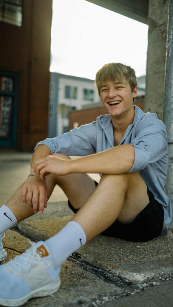 A young man with light brown hair sits on a curb, smiling and looking cheerful. He wears a light blue shirt, black shorts, white socks, and white sneakers. The background shows urban buildings in soft daylight. by Justin Salem Meyer