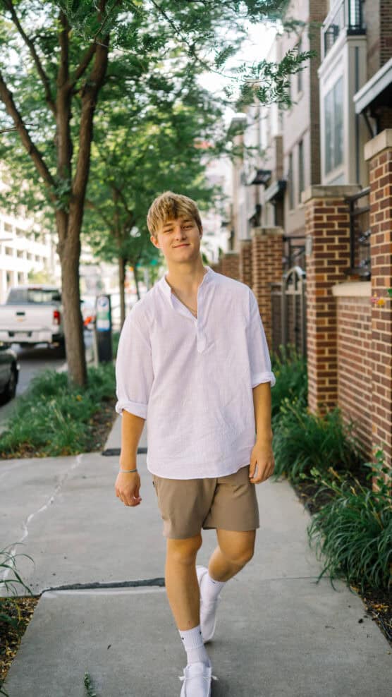A young man wearing a white shirt, khaki shorts, and white sneakers walks on a city sidewalk lined with trees and brick buildings, smiling at the camera. by Justin Salem Meyer