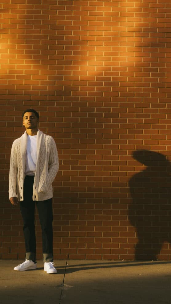 A young person in a cream cardigan, white T-shirt, dark pants, and white sneakers stands against a sunlit red brick wall, casting a long shadow to the right. by Justin Salem Meyer
