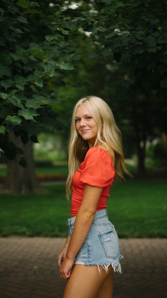 A young woman with long blonde hair, wearing a red top and denim shorts, stands outdoors on a brick path with green trees and grass in the background, smiling slightly and looking at the camera. by Justin Salem Meyer
