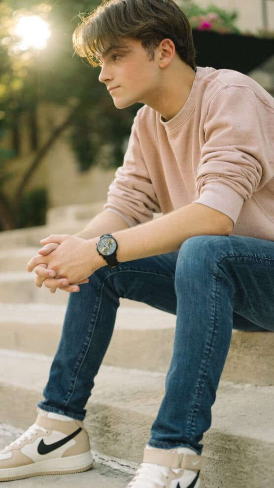 A young man in a light pink sweatshirt, jeans, and beige Nike sneakers sits on outdoor steps, hands clasped, looking thoughtfully to the side as sunlight shines behind him. by Justin Salem Meyer