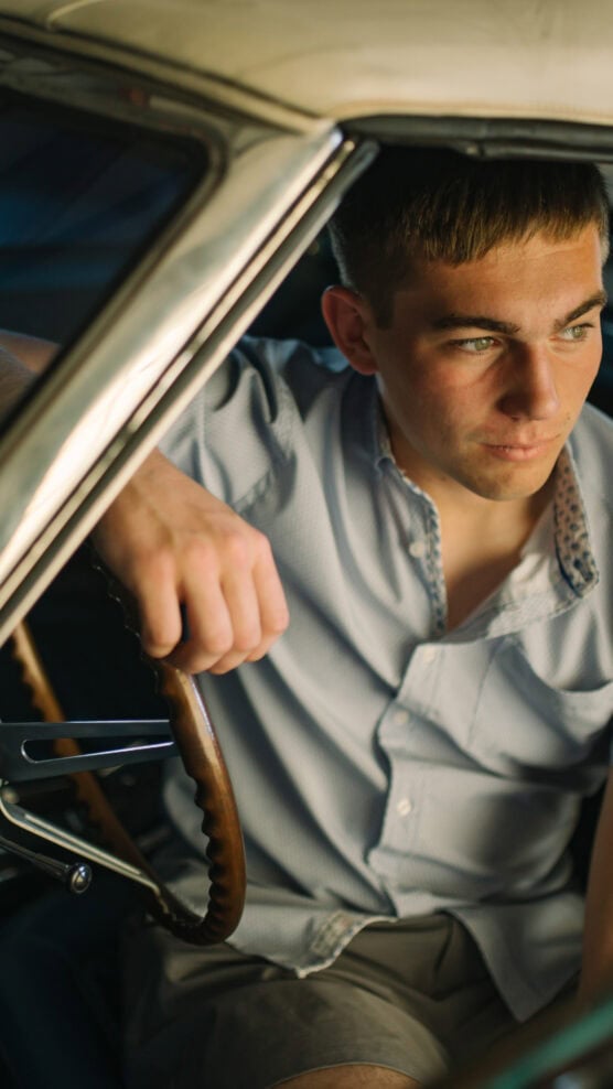A young man with short brown hair sits in the driver’s seat of a classic car, wearing a light blue collared shirt and looking thoughtfully out of the window with one hand on the steering wheel. by Justin Salem Meyer