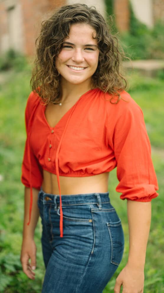 A young woman with curly brown hair wears a bright red crop top and high-waisted blue jeans, smiling outdoors in a green, natural setting with blurred plants and a brick wall in the background. by Justin Salem Meyer