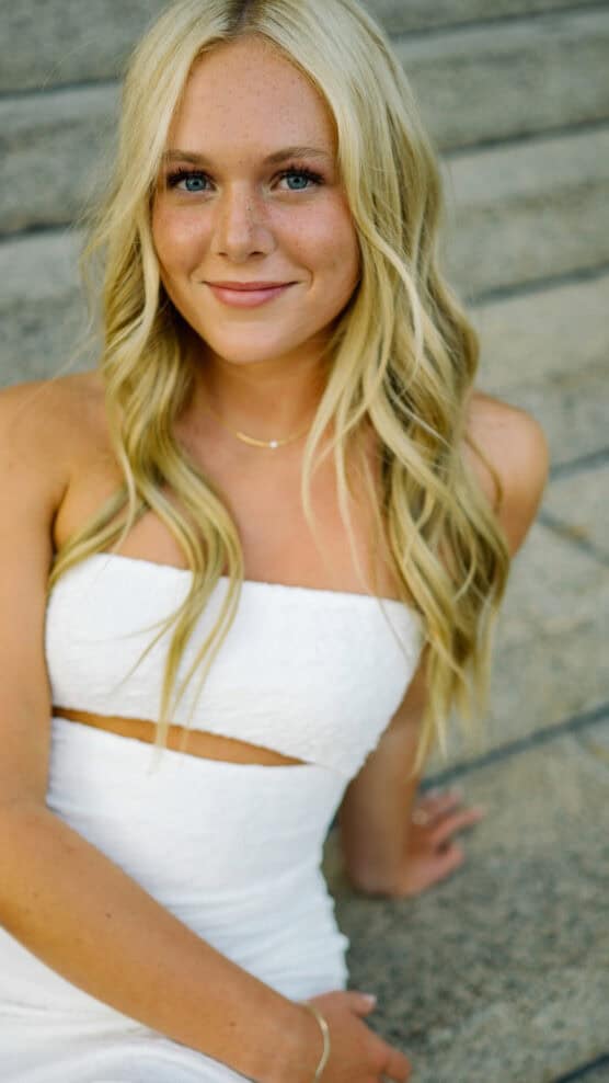 A young woman with long blonde hair and blue eyes sits on outdoor steps, wearing a white strapless dress and a delicate gold necklace, smiling softly at the camera. by Justin Salem Meyer
