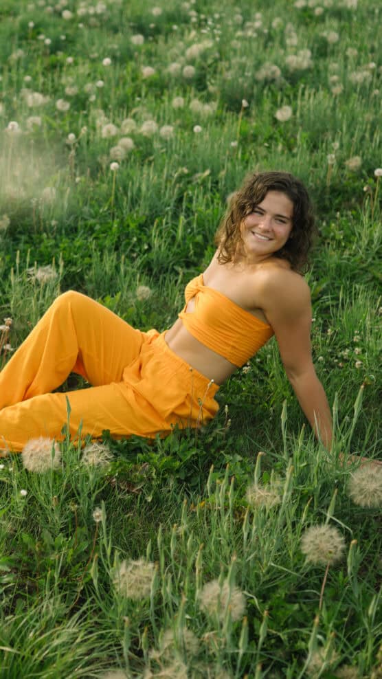 A person with curly hair, wearing a bright orange strapless top and matching pants, sits smiling on green grass in a field dotted with dandelions. by Justin Salem Meyer