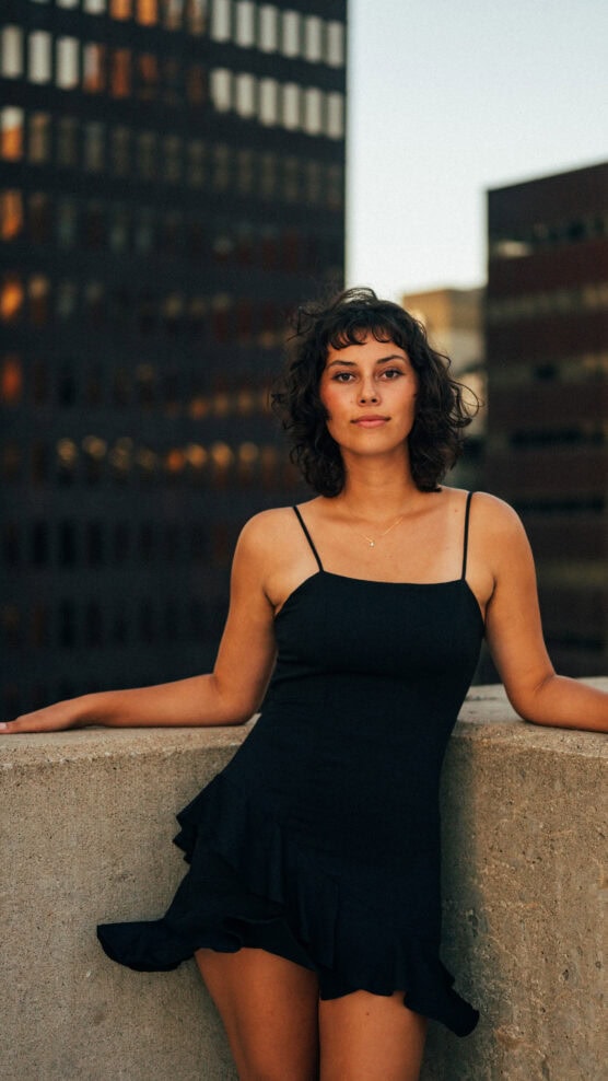A woman with short curly hair in a black dress stands confidently against a concrete ledge on a rooftop, with tall office buildings blurred in the background at sunset. by Justin Salem Meyer