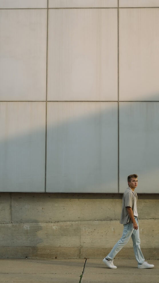 A young man wearing a gray shirt, light jeans, and white sneakers walks along a sidewalk beside a large, pale concrete wall with square panels and a diagonal shadow. by Justin Salem Meyer