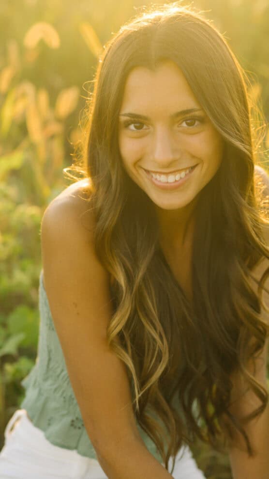 A young woman with long wavy brown hair smiles warmly at the camera while sitting outdoors in a sunlit field, wearing a sleeveless green top and white pants. by Justin Salem Meyer
