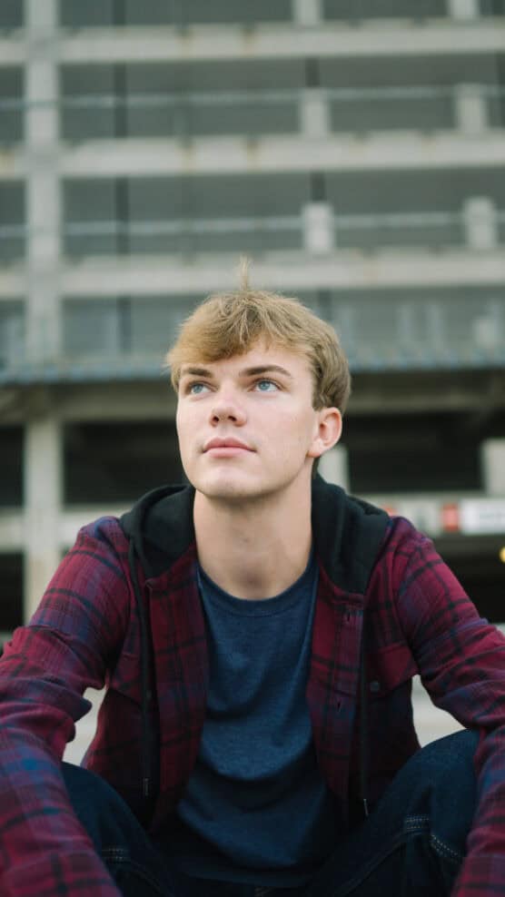 A young man with light brown hair and blue eyes, wearing a red plaid shirt over a dark hoodie, sits outdoors and looks slightly upward. A multi-story concrete building is visible in the blurry background. by Justin Salem Meyer
