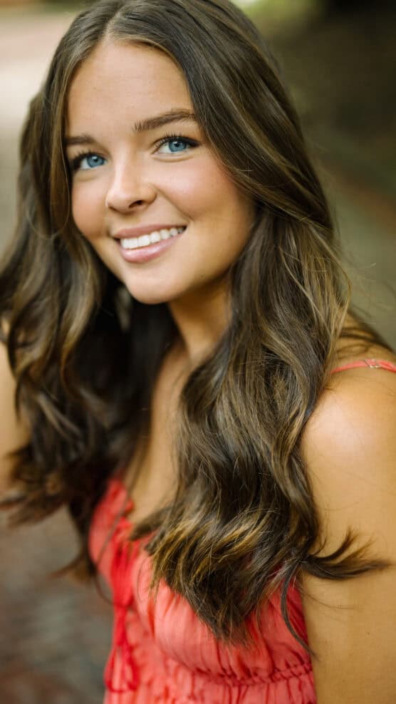 A young woman with long, wavy brown hair and blue eyes smiles at the camera. She is wearing a sleeveless, coral pink top and is outdoors, with a blurred background. by Justin Salem Meyer