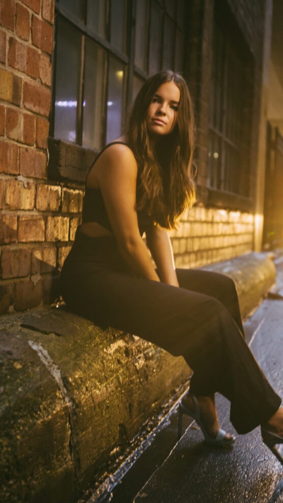 A woman with long brown hair sits on a stone ledge in an urban alleyway at night, wearing a sleeveless black outfit and high heels, with warm lights illuminating the brick wall and wet pavement. by Justin Salem Meyer