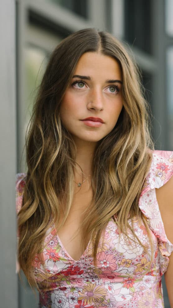 A young woman with long wavy brown hair wearing a floral dress looks thoughtfully to the side while standing near a window. by Justin Salem Meyer