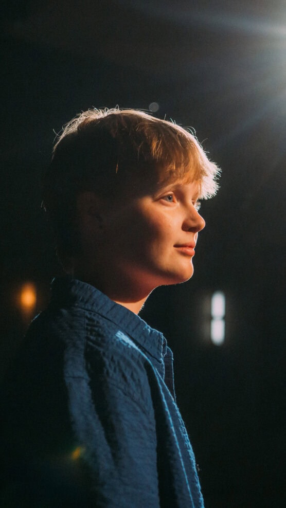 A person with short blonde hair, wearing a textured blue shirt, is shown in profile with soft lighting highlighting their face against a dark, blurred background. by Justin Salem Meyer