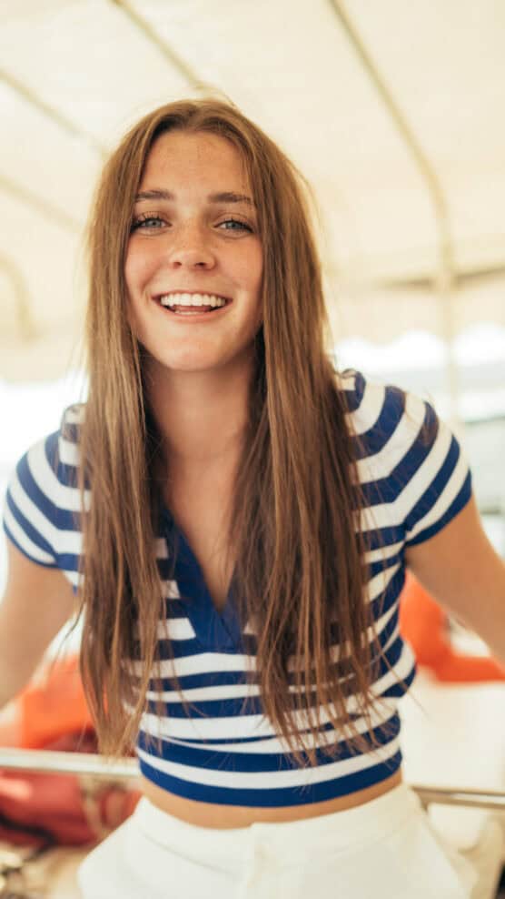 A young woman with long brown hair, wearing a blue and white striped shirt, smiles warmly at the camera in a brightly lit, indoor or sunlit setting. by Justin Salem Meyer