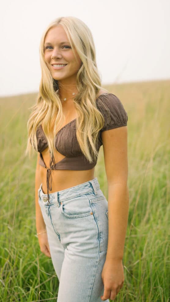 A young woman with long blonde hair, wearing a brown crop top and light blue high-waisted jeans, stands smiling in a grassy field on a sunny day. by Justin Salem Meyer
