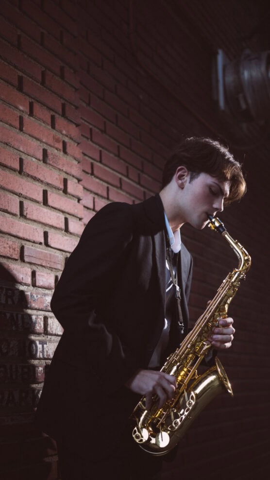 A young person in a dark suit plays a shiny saxophone while standing against a dimly lit brick wall, creating a moody, atmospheric scene. by Justin Salem Meyer
