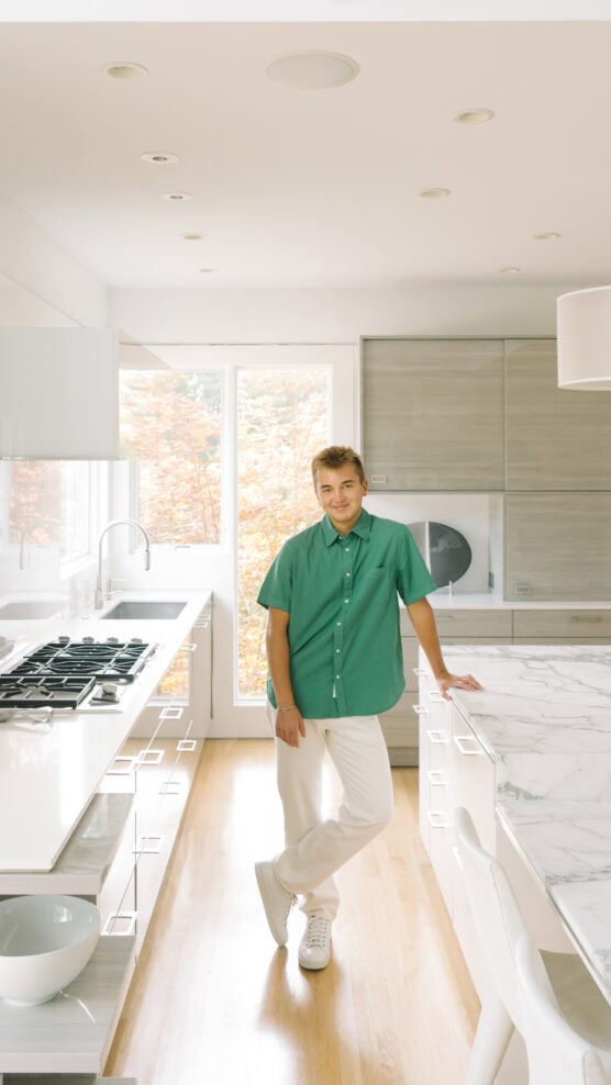 A young person in a green shirt and white pants stands in a modern, bright kitchen with marble countertops, light wood cabinets, and large windows showing autumn trees outside. by Justin Salem Meyer