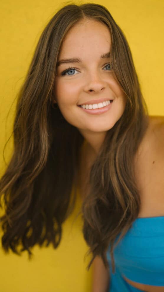 A young woman with long brown hair and blue eyes smiles at the camera against a bright yellow background. She is wearing a blue strapless top. by Justin Salem Meyer
