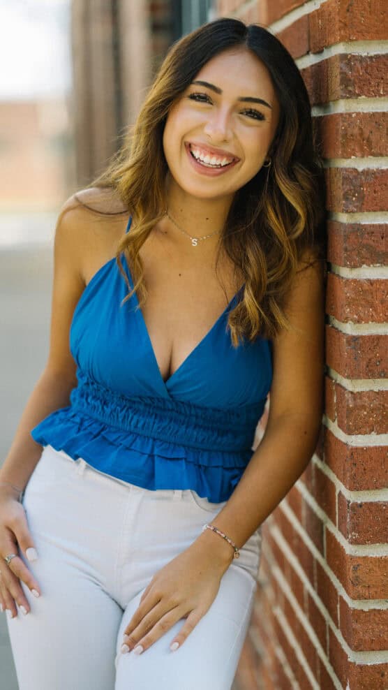 A young woman with long brown hair and a bright smile leans against a brick wall. She is wearing a blue sleeveless top and white pants, with one hand resting on the wall and the other by her side. by Justin Salem Meyer