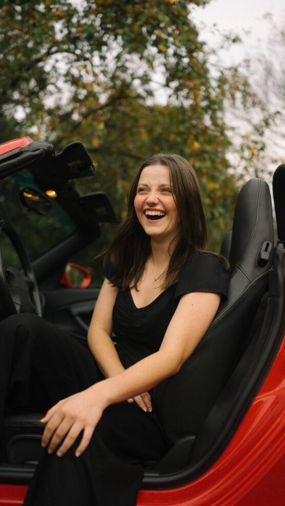 A young woman with long brown hair, dressed in black, sits in the driver’s seat of a red convertible, smiling and laughing with trees in the background. by Justin Salem Meyer