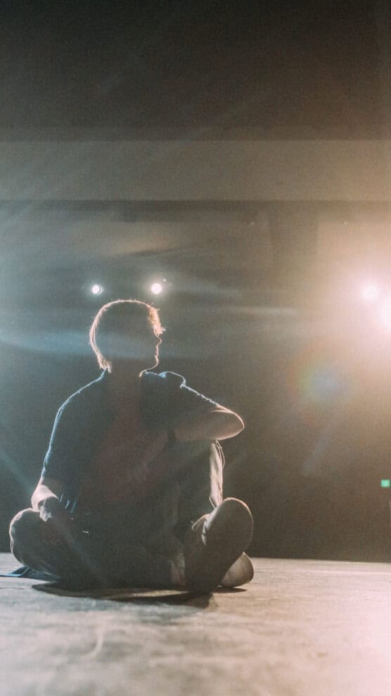 A person sits cross-legged on a stage, illuminated by bright spotlights from behind. Their figure is mostly in shadow, and the scene has a dramatic, contemplative atmosphere. by Justin Salem Meyer
