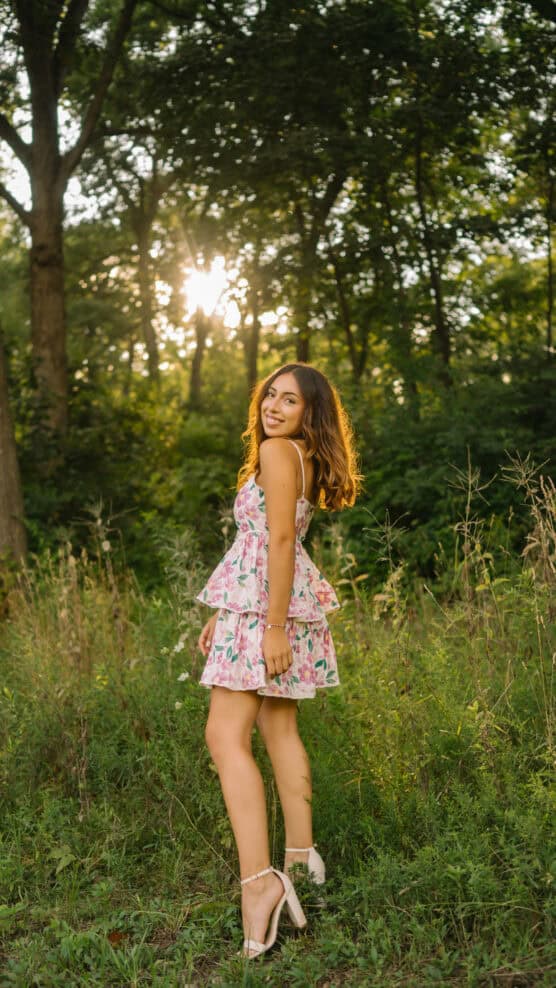 A young woman in a floral dress and heels stands in a grassy, sunlit forest clearing, smiling and looking over her shoulder as sunlight filters through the trees behind her. by Justin Salem Meyer