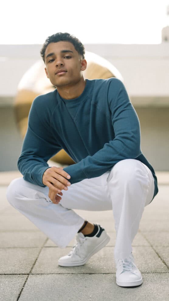 A young man in a teal long-sleeve shirt and white pants squats on a paved surface outdoors, looking confidently at the camera. He is wearing white sneakers, and a blurred sculpture is visible in the background. by Justin Salem Meyer